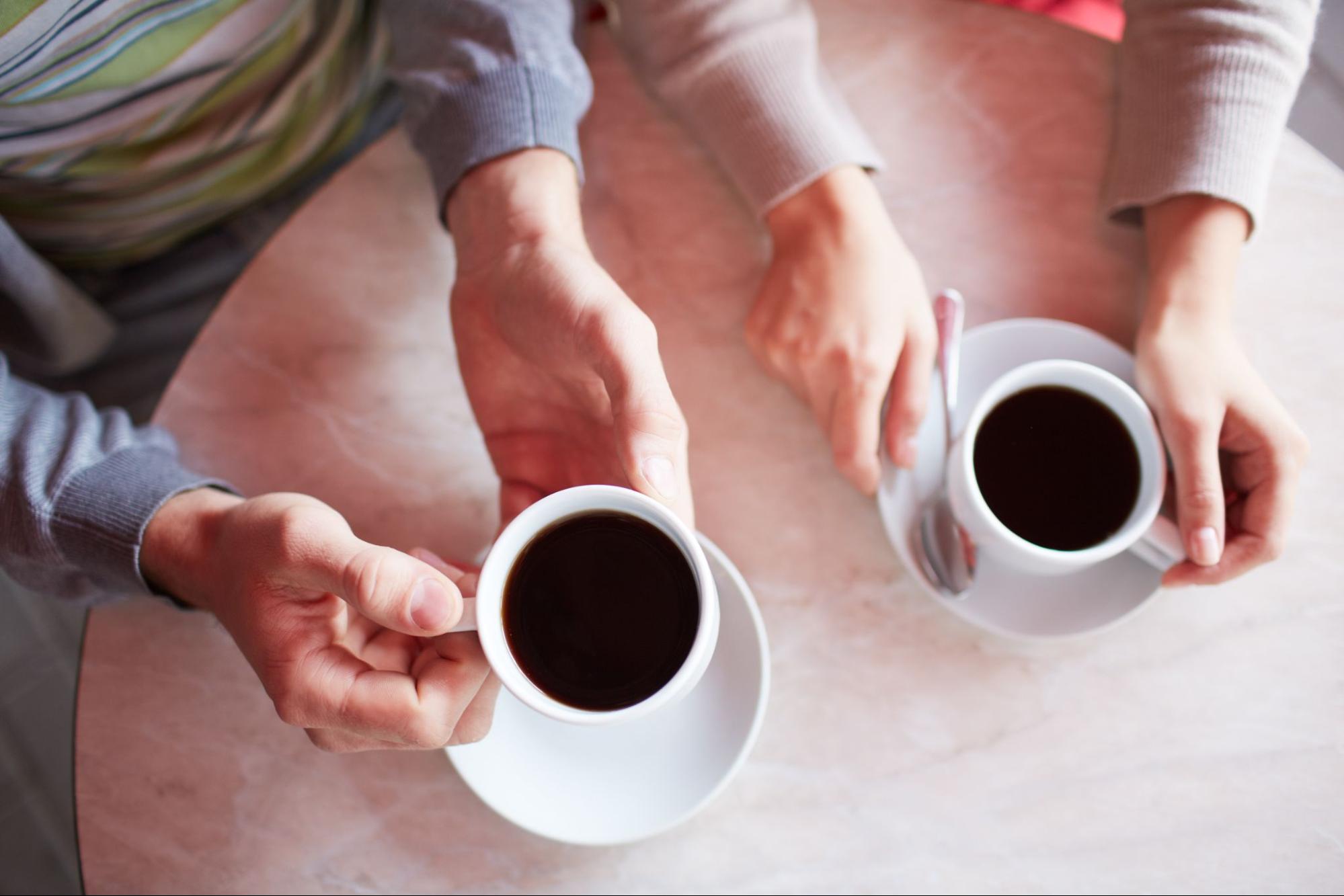 Top-down view of two people at a marble table holding white cups of black coffee.